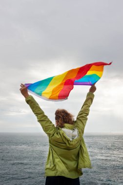 Sırtüstü uzanmış, saçları kıvırcık, kollarını kaldırmış, Brittany, Fransa 'da Pont-Croix Burnu' nun ucunda gökkuşağı bayrağı sallayan, arka planda deniz..
