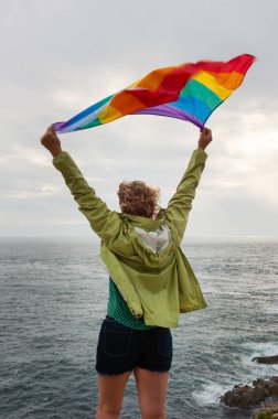 Sırtüstü uzanmış, saçları kıvırcık, kollarını kaldırmış, Brittany, Fransa 'da Pont-Croix Burnu' nun ucunda gökkuşağı bayrağı sallayan, arka planda deniz..