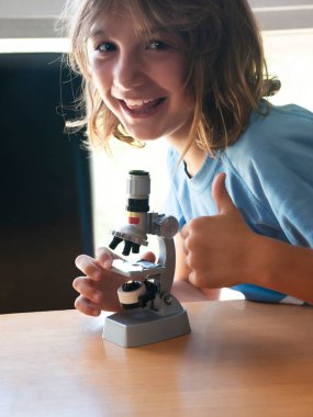 Girl with long brown hair smiling and making the OK sign while working with a school microscope, on a study table with a window in the background.