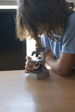 Girl with long brown hair looking at a blood sample through a school microscope on a study table with a window in the background.