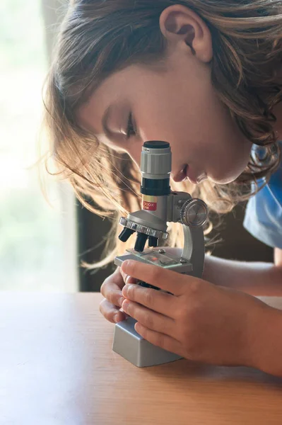 Girl with long brown hair looking at a blood sample through a school microscope on a study table with a window in the background.