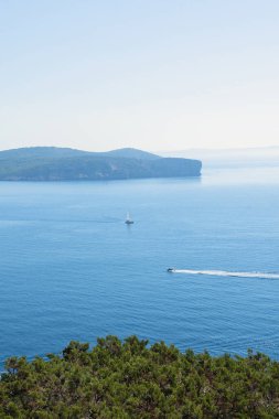 Vertical view from a hill of a cape in Italy with a sailboat in the distance and mountains in the background. Sunny midday with a turquoise Mediterranean Sea on the horizon.