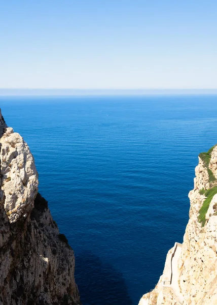 Vertical view of a gorge with cliffs on both sides and a sea inlet. Sunny midday with a turquoise Mediterranean Sea and blue sky on the horizon.