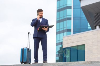 Kendine güvenen bir iş adamı, bir ofis binasının arka planında merdivenlerde duran bir bavulla duruyor.