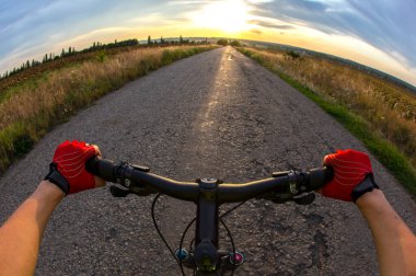 Bisikletçi, elleri bir buğday tarlasının arka planında ve gün batımı gökyüzünde bir bisikletin gidonunu tutuyor. Spor ve seyahat