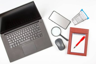Laptop, büyüteç, bilgisayar faresi, akıllı telefon, kalem, not defteri ve beyaz arka planda market sepeti. İş ve mal satın almak için internet üzerinden eşyalar