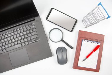 Laptop, büyüteç, bilgisayar faresi, akıllı telefon, kalem, not defteri ve beyaz arka planda market sepeti. İş ve mal satın almak için internet üzerinden eşyalar