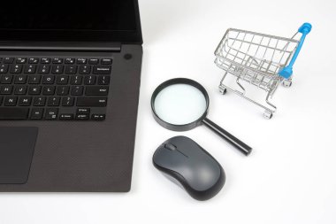 Laptop, büyüteç, bilgisayar faresi ve beyaz arka planda market sepeti. İş ve mal satın almak için internet üzerinden eşyalar