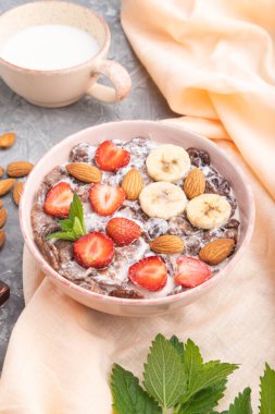 Çikolatalı mısır gevreği, sütlü, çilekli ve bademli gri beton arka planda seramik kase ve turuncu keten tekstil. Yan görünüm, kapat.