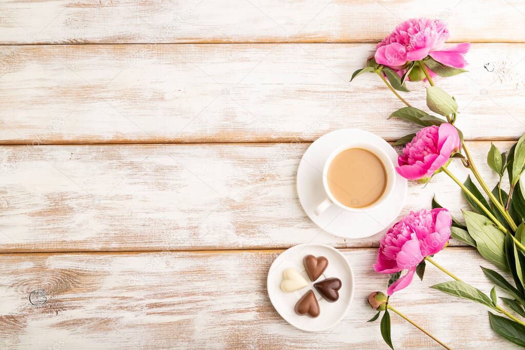 Cup of cioffee with chocolate candies, pink peony flowers on white wooden background. top view, copy space, still life. Breakfast, morning, spring concept.