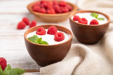 Beyaz ahşap arka plan ve keten tekstil üzerine kil bardaklarda ahududulu yoğurt. Yan görünüm, yakın görünüm, seçici odak.