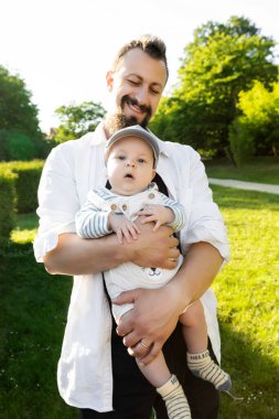 Yeşilliğin arka planında oğlunu kollarına alan bir baba.