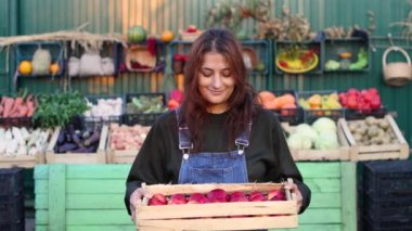 Çiftçi pazarında elmalı kadın çiftçi (satıcı). Sebze ve meyvelerle dolu bir marketin yanında bir kasa elma tutan bir kadının portresi. Elmalara bakar ve gülümser..