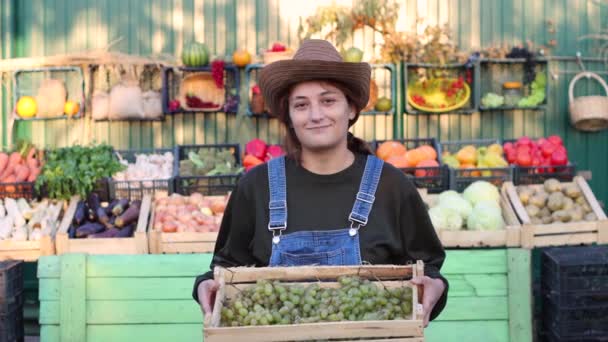 農家の市場でブドウを持つ女性農家 売り手 野菜や果物の市場の屋台の近くにブドウの箱を保持している女性の肖像画 彼女はブドウと笑顔を見て — ストック動画