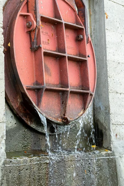 Yuvarlak, kırmızı kapaklı, altından su akan bir kapı. Beton duvara monte edilmiş. 