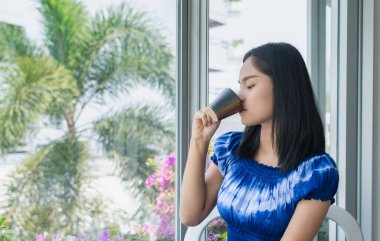 Happy young  Asian woman enjoying and relaxing drinking and looking at a cup of hot coffee drinking coffee the morning sitting by the window in her apartment. Lifestyle concept