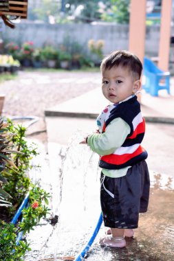 Küçük sevimli Asyalı çocuk güneşli bir günde bahçesinde bahçe çiçeklerini ve çimleri hortumla sularken eğleniyor ve oyun oynuyor. Çocuk küçük yardımcı bahçe işlerini dışarıda öğreniyor. Çocukluk kavramı 