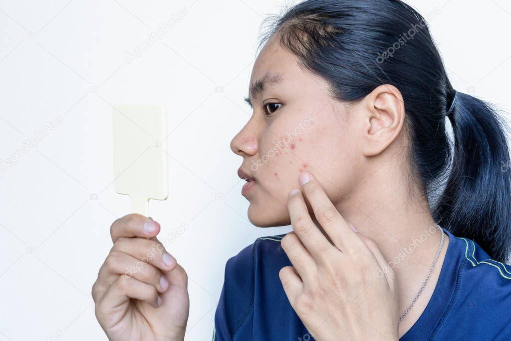 Close-up portrait of a worried young Asian woman in front of small mirror with on white background. Skin problem of pimple, Teenager checking, pointing and touching face by fingers. Skincare concept.
