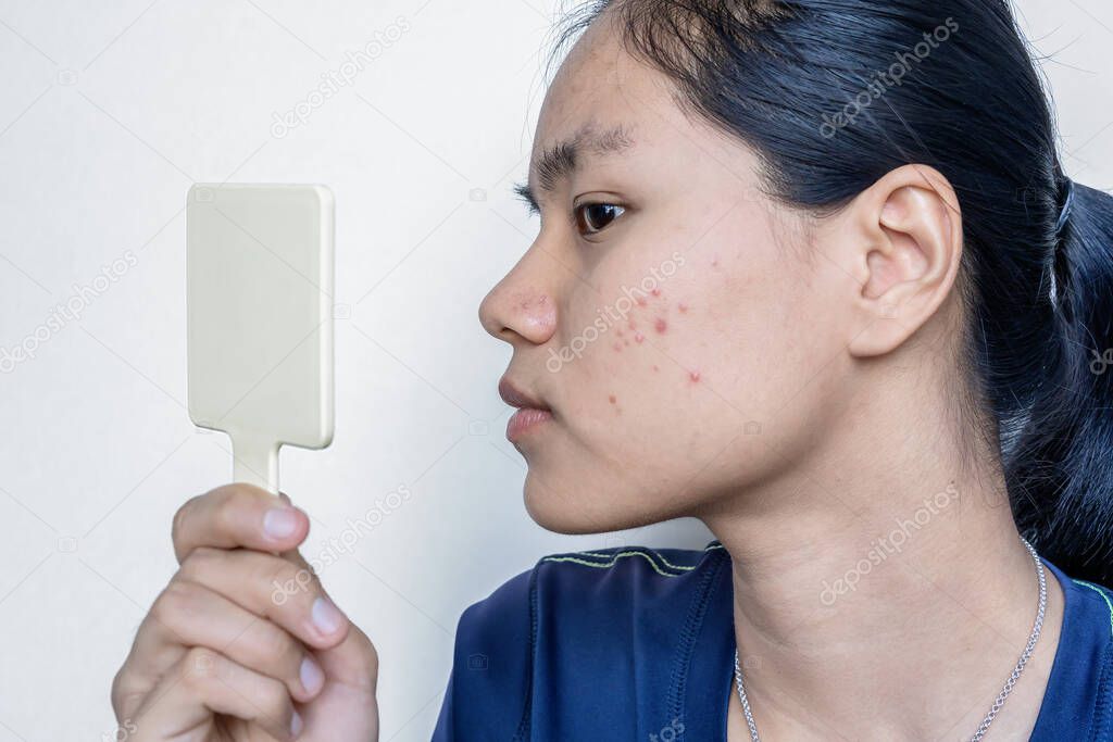 Close-up portrait of a worried young Asian woman in front of the small mirror with on white background. Skin problem of the pimple, Teenager checking her face, problem skincare, and health concept.