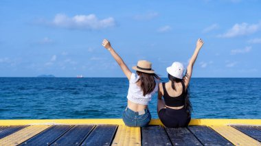 İki mutlu genç kadının el ele tutuşup, tropikal okyanus arka planlarıyla köprüde oturup mavi denizin güzel manzarasının tadını çıkarışının arka görüntüsü. Açık hava yaz yaşam tarzı. Tatiller ve özgürlük kavramı