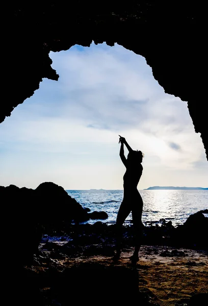 Silhouette of a woman raising hands for yoga or posing to open the mind to beautiful things. Shot of a woman practicing yoga while standing in a cave overlooking the sea at sunrise or sunset.