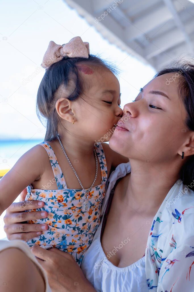 Beautiful young mother laughing with a funny face while her daughter embraces her neck and kisses her cheek outdoor against beach at restaurant or cafe. Happy cheerful family. Mother and baby kissing