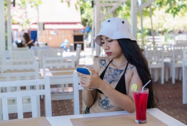 Young beautiful sexy woman in Bikini with fresh juice in restaurant using mobile phone in cafe. Pretty tattooed woman drinking tasty sweet red cocktail, amazing relaxing day on vacation in summertime 