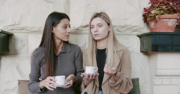 Zwei hübsche Mädchen entspannen sich und unterhalten sich und trinken Kaffee auf der Straße — Stockvideo