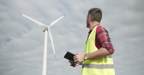 Ervaren ingenieur met tablet kijkt achter op windmolen en kijkt naar camera — Stockvideo