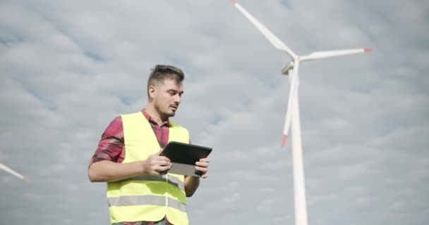 Ervaren ingenieur met tablet kijkt achter op windmolen, werken en glimlachen — Stockvideo