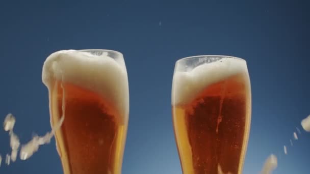 Zwei Gläser voll schäumenden Bieres. sie kollidieren in der Mitte auf blauem Hintergrund — Stockvideo