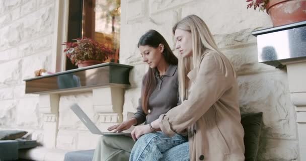 Twee mooie meisjes zitten en werken op een laptop, vlakbij een terras — Stockvideo