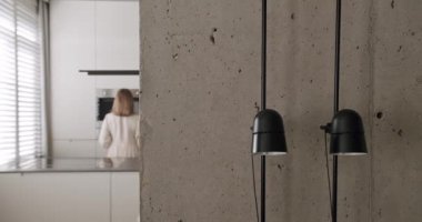 Bir kadın minimalist bir iç mekanda kahve yapar, lambaya odaklanır. 