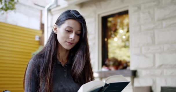 Mujer joven sonriente está leyendo interesante libro al aire libre, hojeando a través de un libro — Vídeo de stock