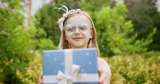 Красивая 7-летняя девочка в очках, дарит подарок в синей коробке с луком. — стоковое видео