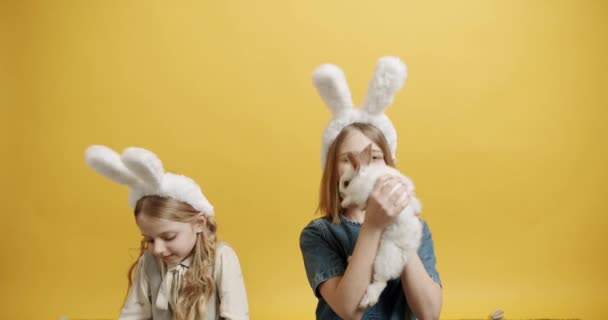 Симпатичные девочки с кроличьими ушами, играющие с кроликом на изолированном фоне — стоковое видео