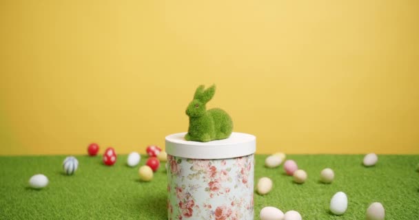Juguete de conejo sobre un fondo amarillo de hierba verde con muchos huevos de colores, Texto. — Vídeo de stock