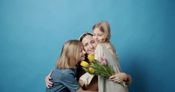 Mamãe abraços Filhas e beijos, com flores, posando em um fundo azul — Vídeo de Stock