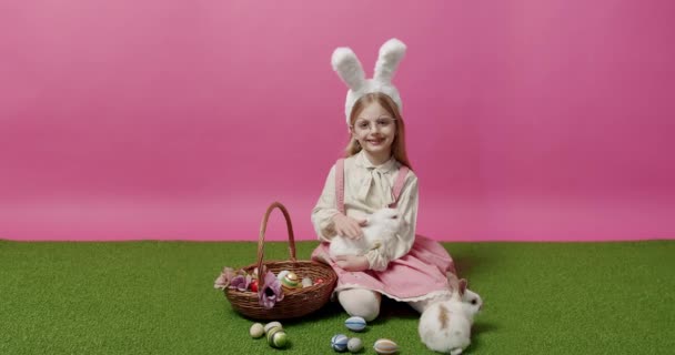 Маленька дівчинка з вухами кролика тримає кошик з кроликом в руці, позує, посміхається — стокове відео