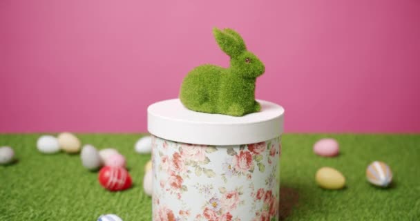Игрушка кролика на розовом фоне зеленой травы со многими цветными яйцами, текст. — стоковое видео