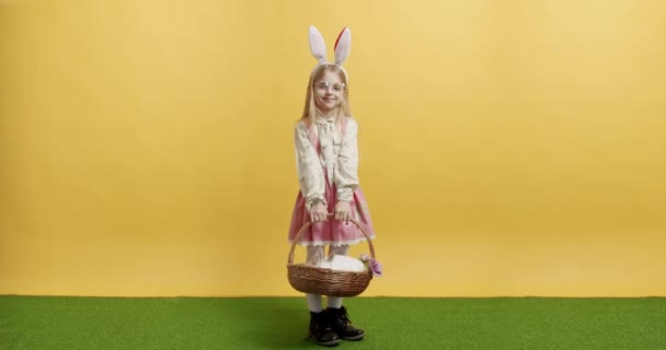 Pembe elbiseli küçük kız elinde tavşanla bir sepet tutuyor, poz veriyor, gülümsüyor. — Stok video