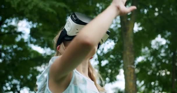 Ένα έφηβο κορίτσι φοράει γυαλιά εικονικής πραγματικότητας και παίζει προσομοίωση 3D βιντεοπαιχνίδι στη φύση. — Αρχείο Βίντεο