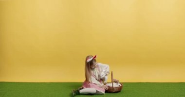 Pembe elbiseli küçük kız elinde tavşanla bir sepet tutuyor, poz veriyor, gülümsüyor.