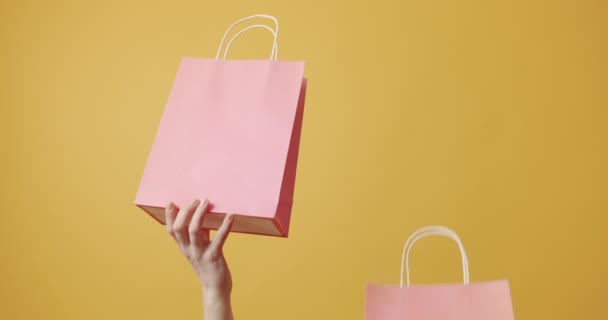 Vrouwen handen tonen roze papieren boodschappentassen op een geïsoleerde gele achtergrond. — Stockvideo