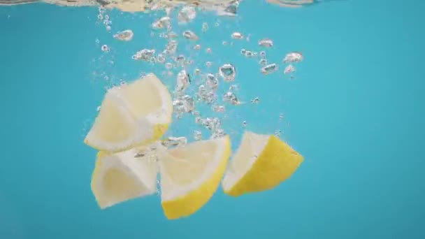 Αργή κίνηση της πτώσης φέτες λεμονιού σε νερό πιτσιλίσματος σε μπλε φόντο — Αρχείο Βίντεο
