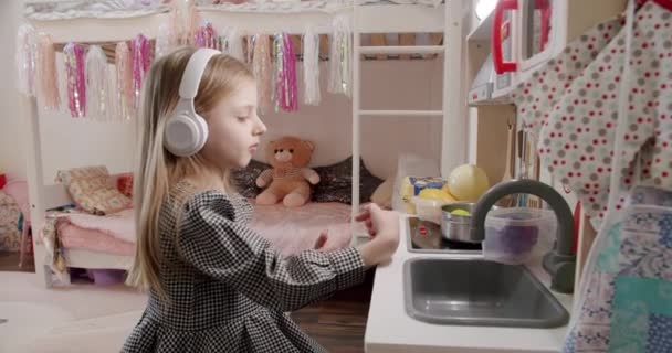 A menina de 7 anos está brincando no quarto das crianças com fones de ouvido brancos. — Vídeo de Stock