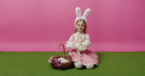 Tavşan kulaklı küçük kız elinde tavşanla bir sepet tutuyor, poz veriyor, gülümsüyor. — Stok video