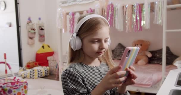 A menina de 7 anos na sala das crianças com fones de ouvido e usando smartphone. — Vídeo de Stock