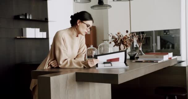 Seriöst avslappnad kvinna läser bok på moderna hem i mysiga kök interiör. — Stockvideo