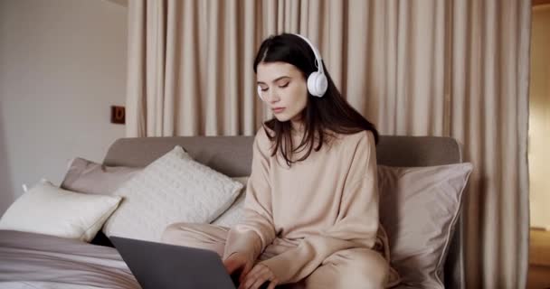 Молодая красивая женщина с ноутбуком с беспроводными наушниками, сидя в уютной кровати — стоковое видео